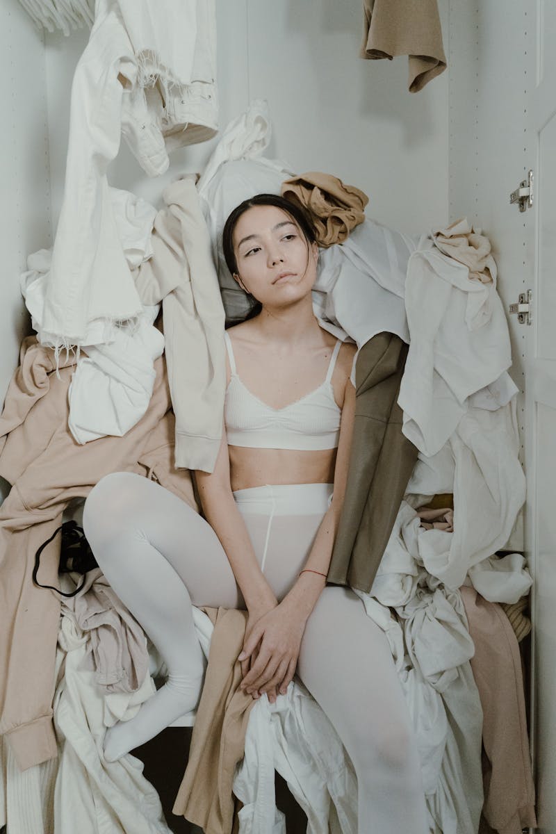 Asian woman sits in a messy wardrobe, symbolizing consumerism and clutter at home.